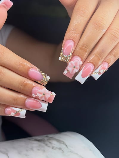 A close-up shows hands with long square nails featuring a pink French ombre, intricate gold rhinestones, and delicate 3D pink and white floral accents.