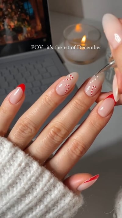 A hand with glossy, almond-shaped nails featuring a sheer nude base; some nails have bright red French tips, while others display delicate red and white candy c