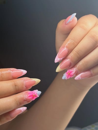 Close-up of stiletto nails with clear bases, pastel pink, blue, and yellow French tips, some adorned with white dots, and delicate 3D pink flowers with gold bea
