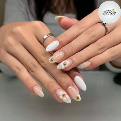 Close-up of a pair of hands displaying almond-shaped nails with a mix of sheer nude and solid white polish, featuring 3D white floral accents with gold centers