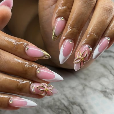 Close-up of long stiletto nails featuring a sheer pink base, white V-shaped French tips, gold metallic accents, 3D gold flowers, pearls, and rhinestones.
