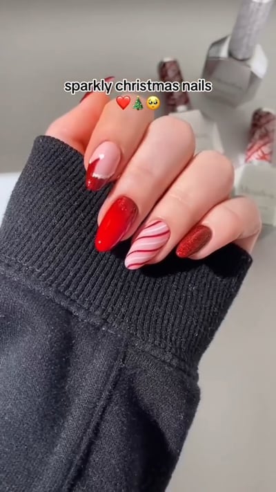 A hand displays a set of sparkly, almond-shaped Christmas nails featuring red glitter, a red French tip, solid red, and a nude nail with red candy cane stripes.