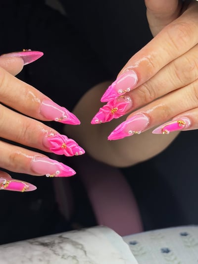 A close-up shows long, sharp stiletto nails with a pink ombre, adorned with vibrant 3D pink flowers, gold beads, and white pearls.