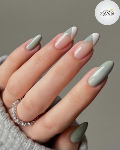 A hand with light skin tone displays medium-length almond nails, featuring a mix of solid sage green and geometric French tips in nude, sage green, and white.