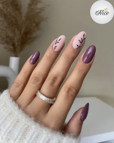 A hand displays oval-shaped nails featuring a mix of glossy deep mauve and matte light pink with black leaf designs.