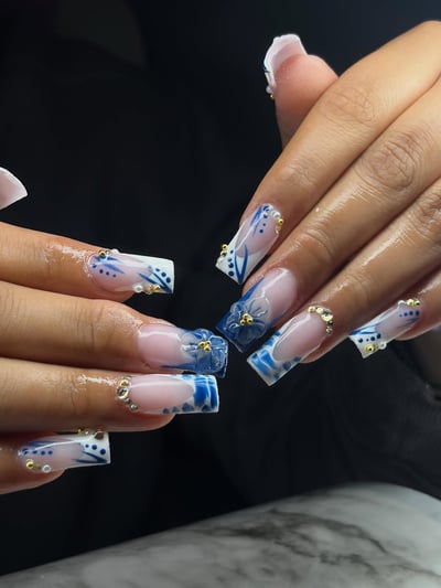 A close-up of long, glossy square nails featuring intricate blue and white designs, 3D flowers, gold studs, and rhinestones.