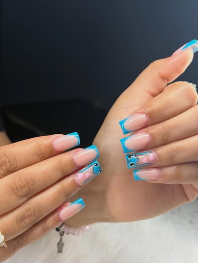 A close-up of two hands displaying long, glossy, soft square nails with vibrant blue French tips and playful blue cartoon character art accented with pink heart