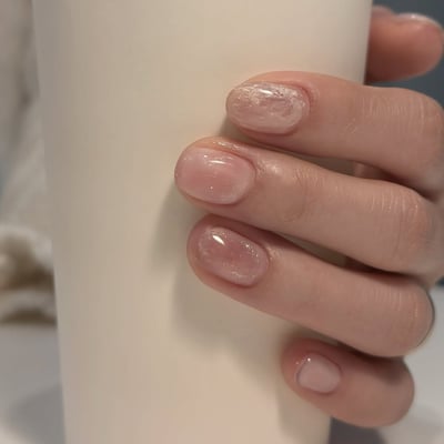 Short, oval nails feature a sheer pink base with subtle white marble patterns and a delicate glitter finish.