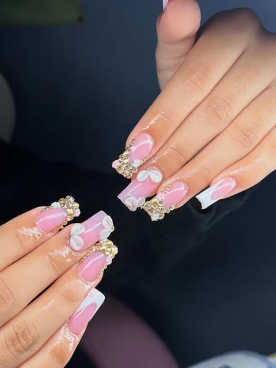 Close-up of hands displaying long, glossy pink and white nails adorned with abundant gold rhinestones and delicate 3D white bows.