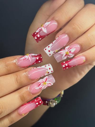Close-up of long, square acrylic nails featuring a pink base with red and white polka dot French tips, adorned with intricate 3D white and red flowers and gold