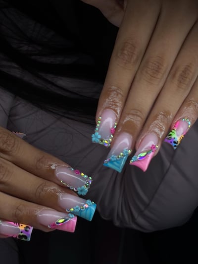 Long square nails feature clear bases with colorful pink and blue tips, iridescent rhinestones, 3D blue flowers, and a vibrant animal print.