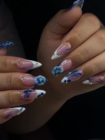 A close-up shows long, pointed almond nails with sheer pink bases, white tips, intricate blue floral patterns, 3D blue flowers, and gold stud accents.