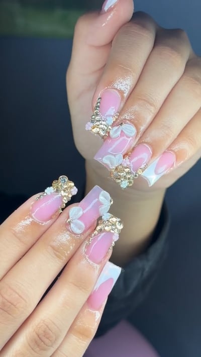A hand displays long, square-shaped nails with a pink base, white French tips, 3D white bows, and elaborate gold and metallic rhinestone embellishments.