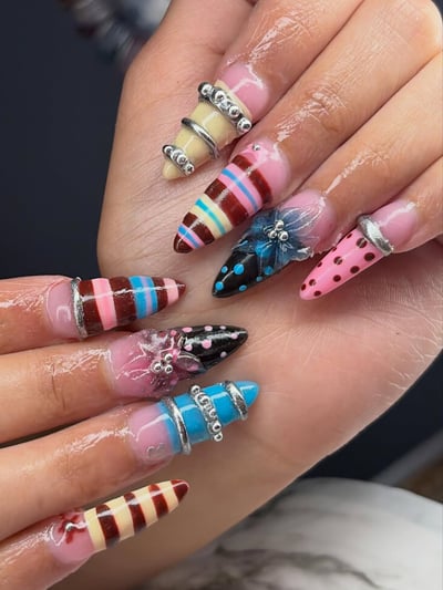 Extreme stiletto nails with vibrant pink, black, cream, brown, blue designs, featuring stripes, polka dots, 3D silver rings, and floral accents.