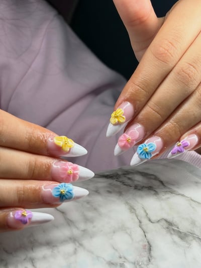 Close-up of hands with long, pointed almond nails featuring a French tip design adorned with various pastel 3D flowers with gold centers.