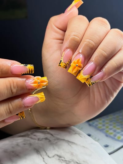 A close-up of long, tapered square nails featuring sheer pink bases, vibrant yellow and black tiger print French tips, and 3D orange flowers with rhinestone acc