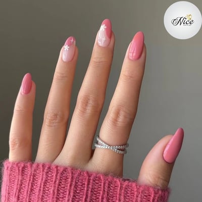 A hand with medium-length, glossy pink almond nails, featuring two accent nails with pink tips, nude bases, and delicate white floral designs.
