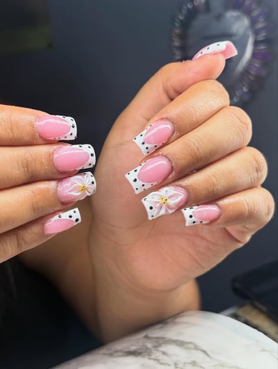 A hand displays long, glossy square nails featuring a pink base, white French tips with black polka dots, and delicate 3D white flowers with gold centers.