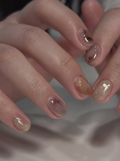 Oval, medium-length nails with translucent nude marble design, featuring brown swirls and gold foil accents.