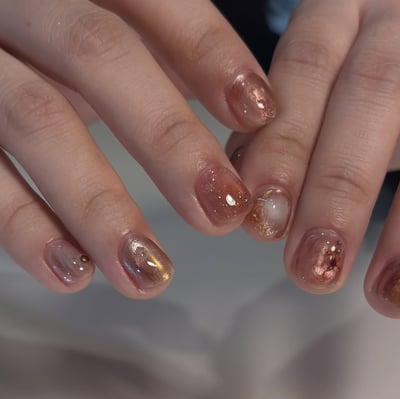 Medium oval nails feature a translucent reddish-brown cat-eye effect with gold foil accents and a small gold stud.