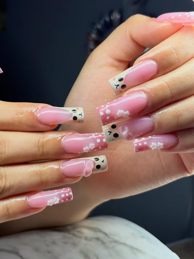 Close-up of hands displaying long, glossy square nails with pink bases and French tips featuring cute cartoon faces, white polka dots, and raised 3D white flowe