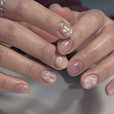 Oval, medium-length nails with a mix of sheer nude, dusty rose, and milky white polishes, adorned with glitter, gold foil, and 3D shell charms.