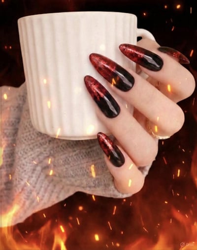 A hand holds a white mug, showcasing long, almond-shaped nails with a glittery red-to-black ombre design, set against a fiery background.