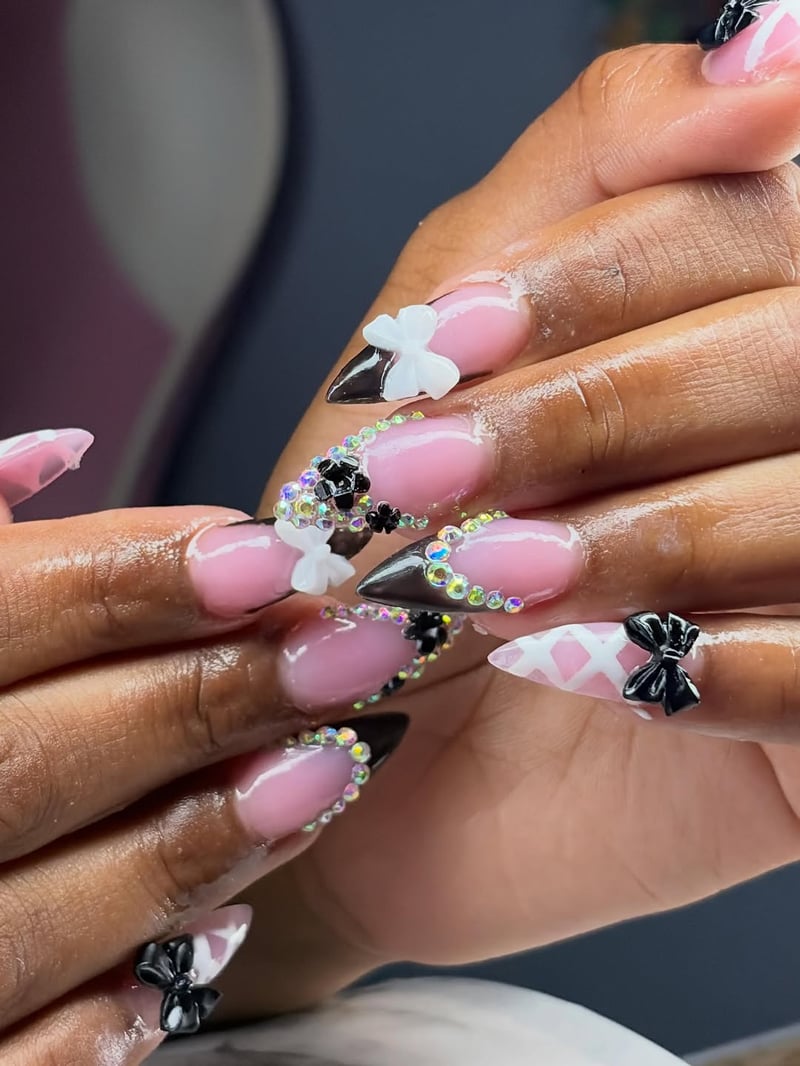 Close-up of long, pointed pink nails with black tips, white and black 3D bows, and iridescent rhinestone accents, worn by a person with deep skin tone.