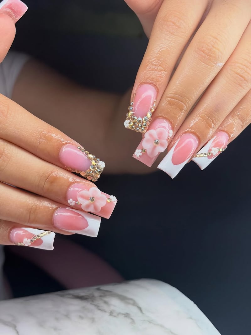 A close-up shows hands with long square nails featuring a pink French ombre, intricate gold rhinestones, and delicate 3D pink and white floral accents.
