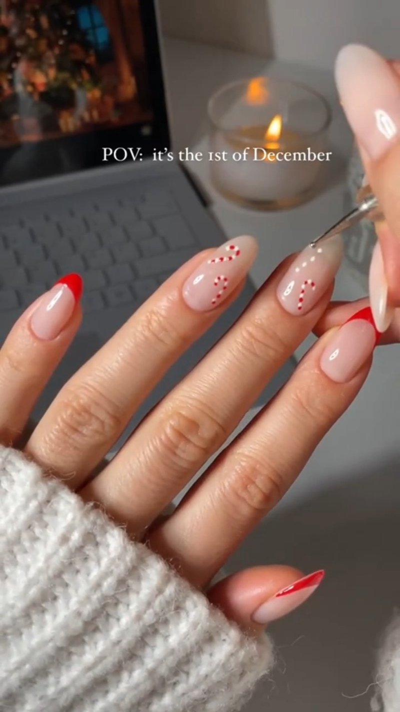 A hand with glossy, almond-shaped nails featuring a sheer nude base; some nails have bright red French tips, while others display delicate red and white candy c