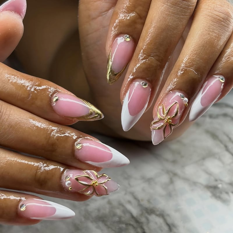 Close-up of long stiletto nails featuring a sheer pink base, white V-shaped French tips, gold metallic accents, 3D gold flowers, pearls, and rhinestones.