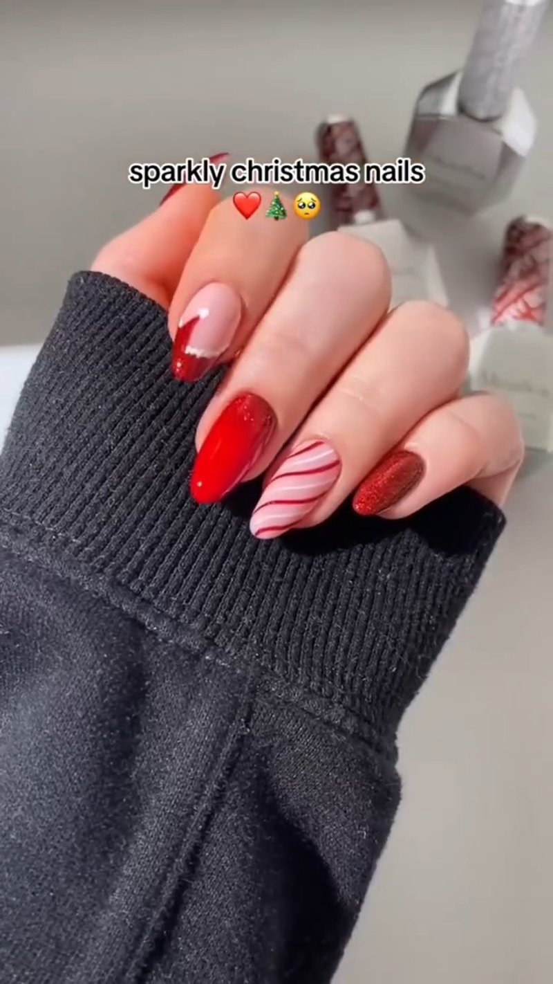 A hand displays a set of sparkly, almond-shaped Christmas nails featuring red glitter, a red French tip, solid red, and a nude nail with red candy cane stripes.