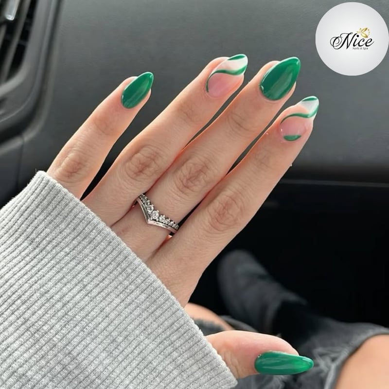 A close-up of a hand with almond-shaped nails featuring a glossy emerald green polish and accent nails with nude bases and elegant green and white swirl designs