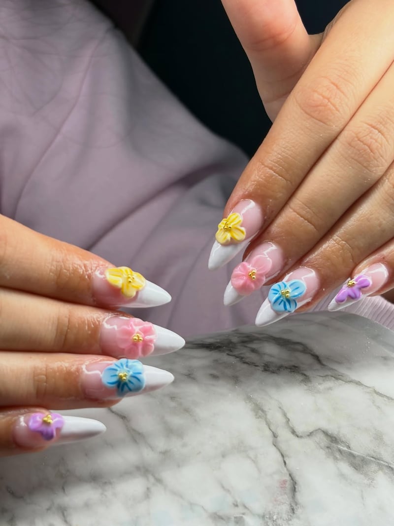 Close-up of hands with long, pointed almond nails featuring a French tip design adorned with various pastel 3D flowers with gold centers.