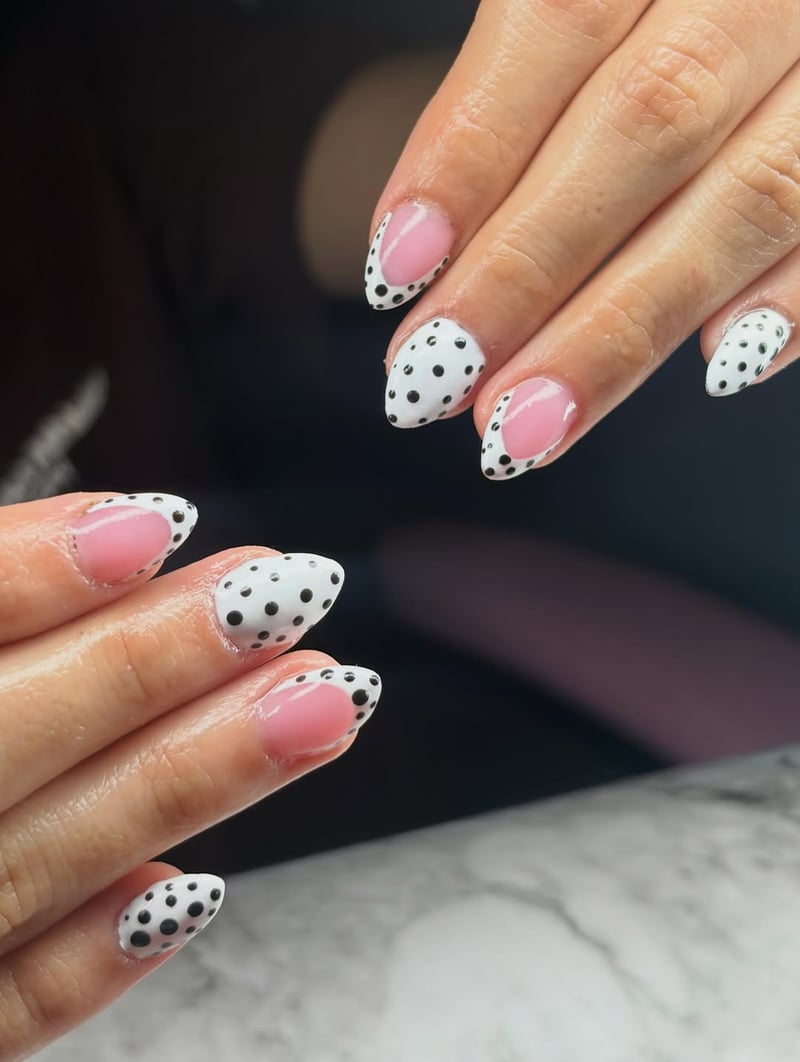 Close-up of hands displaying almond-shaped nails with sheer pink bases and white tips featuring black polka dots in a modern French design.