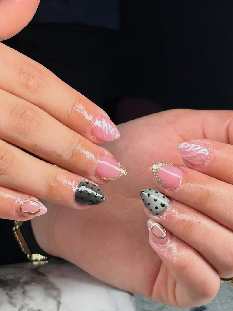 A close-up of hands displaying a mixed nail art design featuring pink nails with gold glitter tips and white swirls, alongside black nails with white polka dots