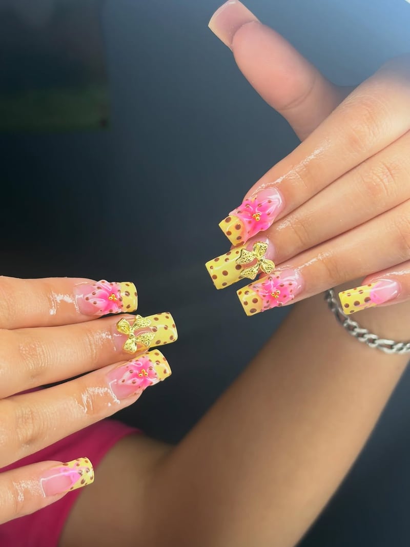 A close-up of hands displaying long, coffin-shaped nails with clear bases, yellow tips featuring brown polka dots, 3D pink flowers, and gold bow charms.